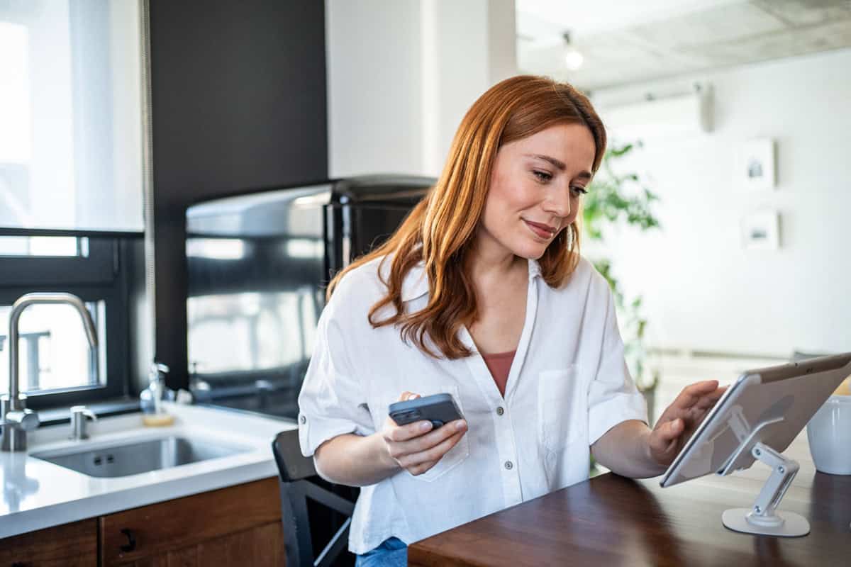 Woman using a tablet at a kitchen table, holding a smartphone and smiling.